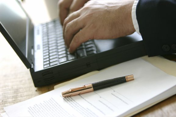 Person tippt auf einer Laptop-Tastatur, neben dem Laptop liegt ein Kugelschreiber und Papiere.