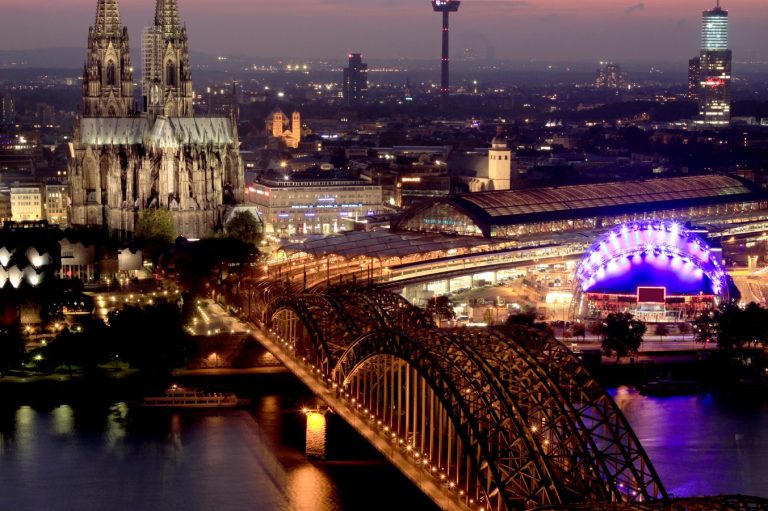 Kölner Dom und Hohenzollernbrücke bei Abenddämmerung mit beleuchteten Unterbrechungen.