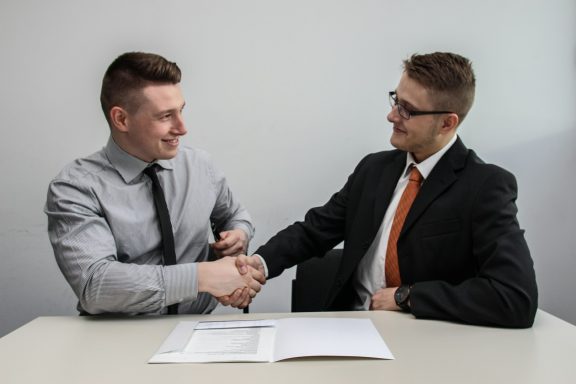Zwei Männer schütteln sich die Hände am Tisch, Dokumente liegen daneben.