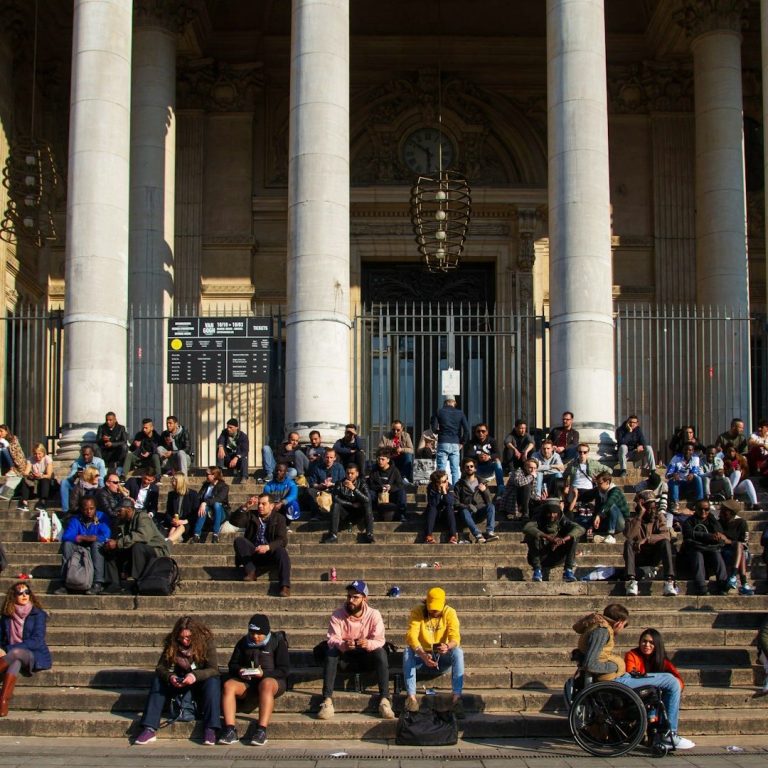Menschen sitzen auf Treppen vor einem großen Gebäude, einige in Rollstühlen.