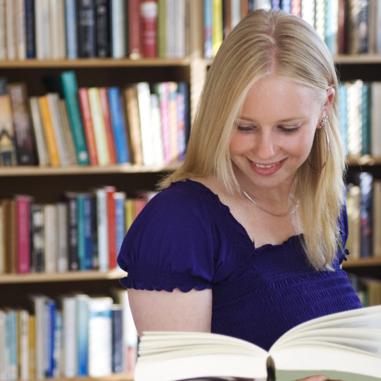Eine lächelnde Frau liest ein Buch vor einem Bücherregal.
