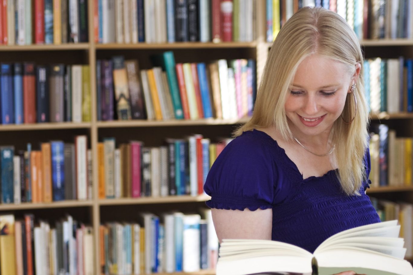 Frau mit langen blonden Haaren, die lächelnd in einem Buch blättert, vor einem Bücherregal.