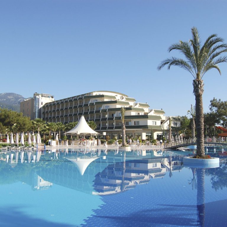 Schwimmbecken mit Palmen und Hotelgebäude im Hintergrund unter blauem Himmel.