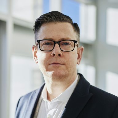 Mann mit Brille, blonden Haar und formeller Kleidung, steht vor einem hellen Hintergrund.