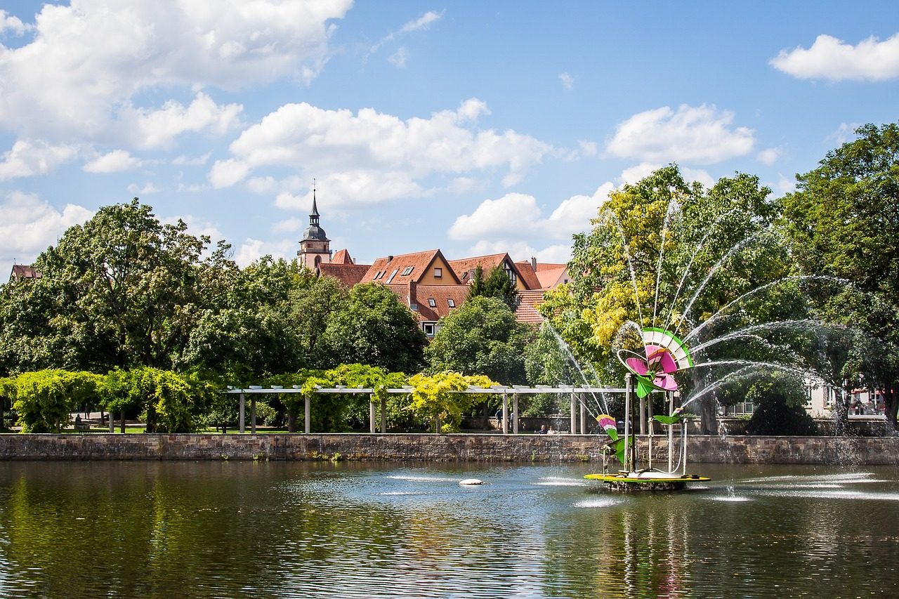 Grüner Park mit einem Teich, Springbrunnen und blühenden Pflanzen im Hintergrund.