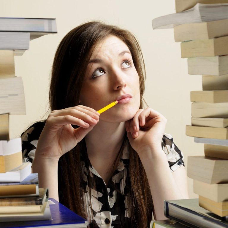 Frau mit Bleistift in der Hand, blickt nachdenklich zwischen Büchertürmen.