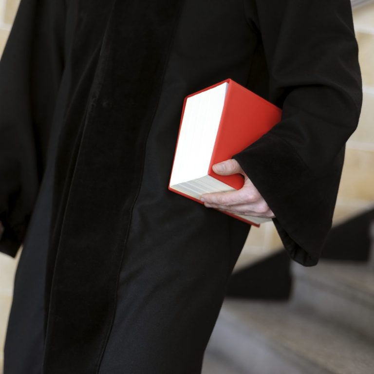 Eine Person in Robe hält ein rotes Buch mit weißem Rücken.