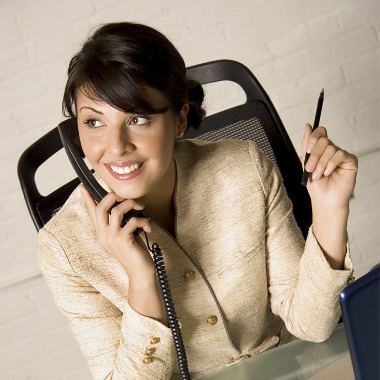 Frau mit Telefon und Stift, lächelnd in einem Büroumfeld.