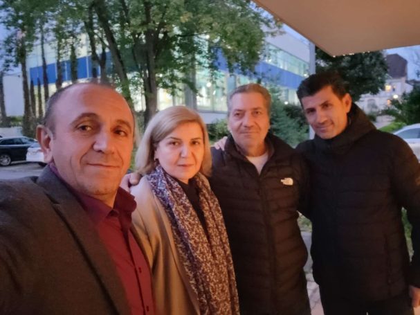 Vier Personen stehen zusammen im Freien, umgeben von Bäumen und einem Gebäude im Hintergrund.