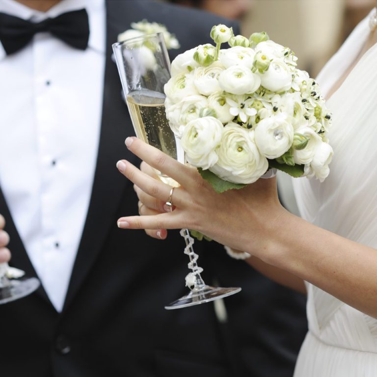 Braut hält ein Blumengebinde und ein Champagnerglas, Bräutigam daneben.