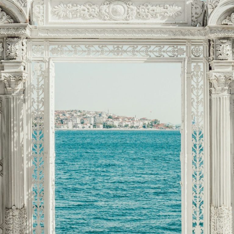 Blick durch einen kunstvoll verzierten Rahmen auf ruhiges, blaues Wasser und eine Küstenstadt.