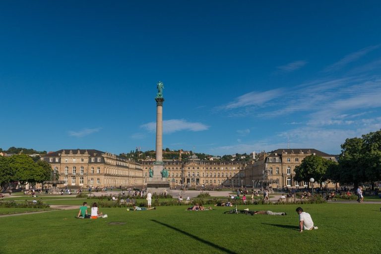 Blick auf die rotunde Säule mit der Statue, umgeben von Grünflächen und historischem Gebäude.