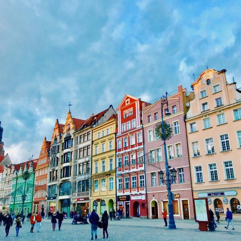 Bunte Häuserfassaden am Platz mit Menschen und bewölktem Himmel.