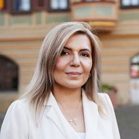 Frau mit langen, blonden Haaren in einem hellen Blazer vor einem historischen Gebäude.
