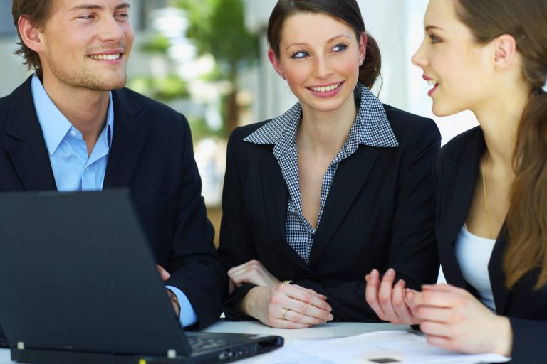 Drei Personen in Anzügen diskutieren lebhaft am Tisch mit Laptop.