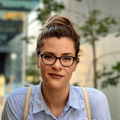 Junge Frau mit Brille und hochgestecktem Haar, lächelt vor urbanem Hintergrund.