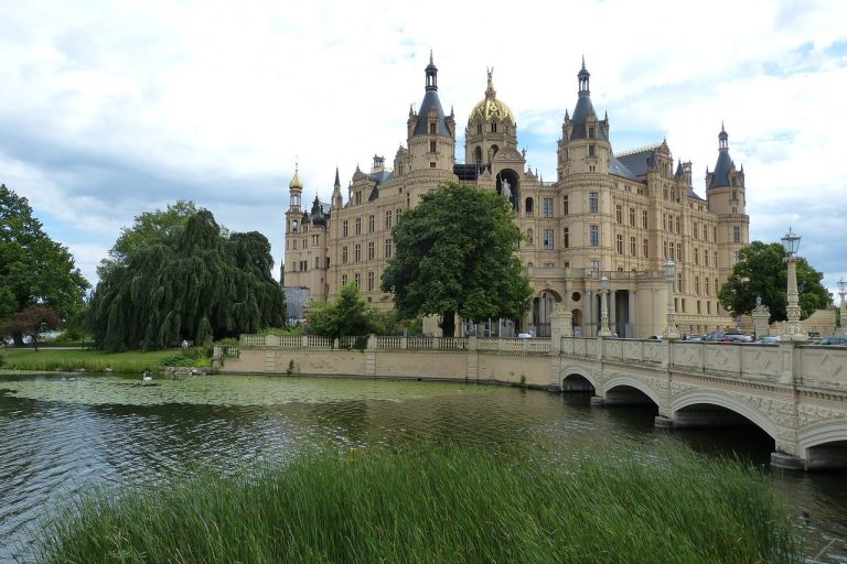Schloss Schwerin mit Brücke und See, umgeben von Bäumen und bewölktem Himmel.