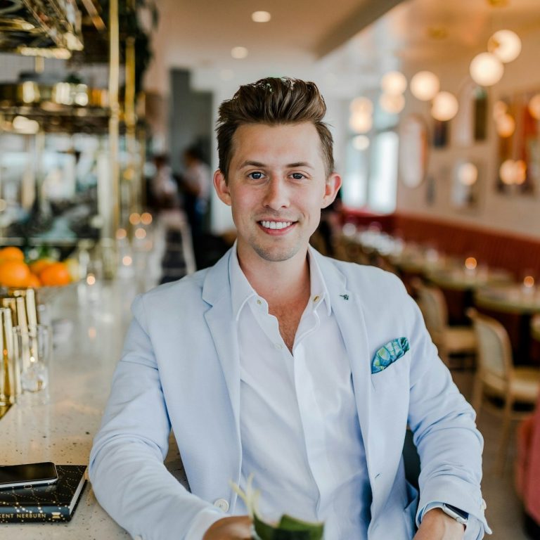 Ein lächelnder Mann in einem eleganten Outfit sitzt an einer Bar mit Getränk.