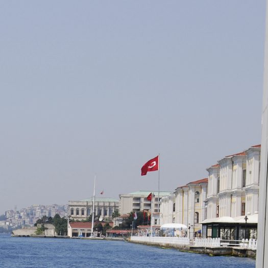 Helle Uferlinie mit weißem Gebäude und der rot-weißen Flagge im Hintergrund.