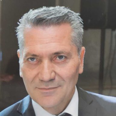 Mann mit grauen Haaren in einem Anzug, neutraler Gesichtsausdruck.